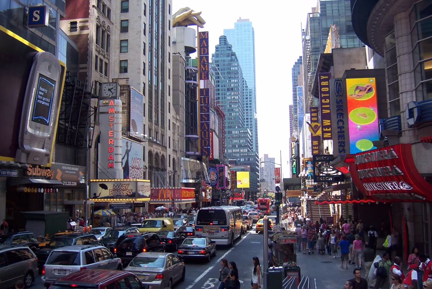 A street with two lanes of mixed-traffic in either direction, and third bus/taxi lane. Pedestrians are squashed into the fairly narrow pavements on each side, with a few waiting to cross the backed-up lanes of traffic. The cars undoubtedly dominate this street, despite the many many shops, restaurants and attractions on it, and even a subway station on the left side.